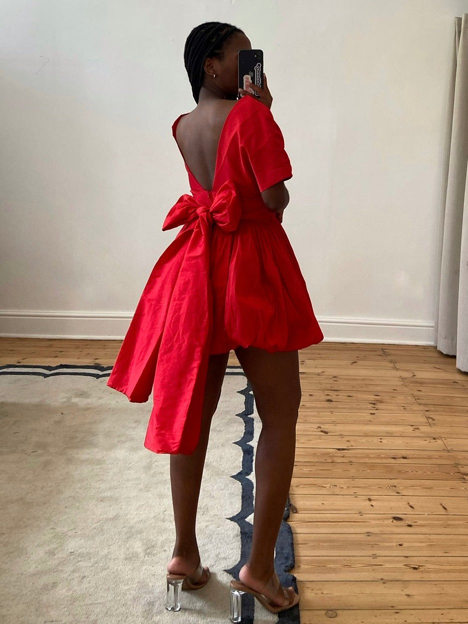 The Stella Mini by Kat Van Duinen – a red mini dress with a bubble hem skirt and tie waist belt, perfect for cocktail parties and festive events. Bold, chic, and available now for rental.