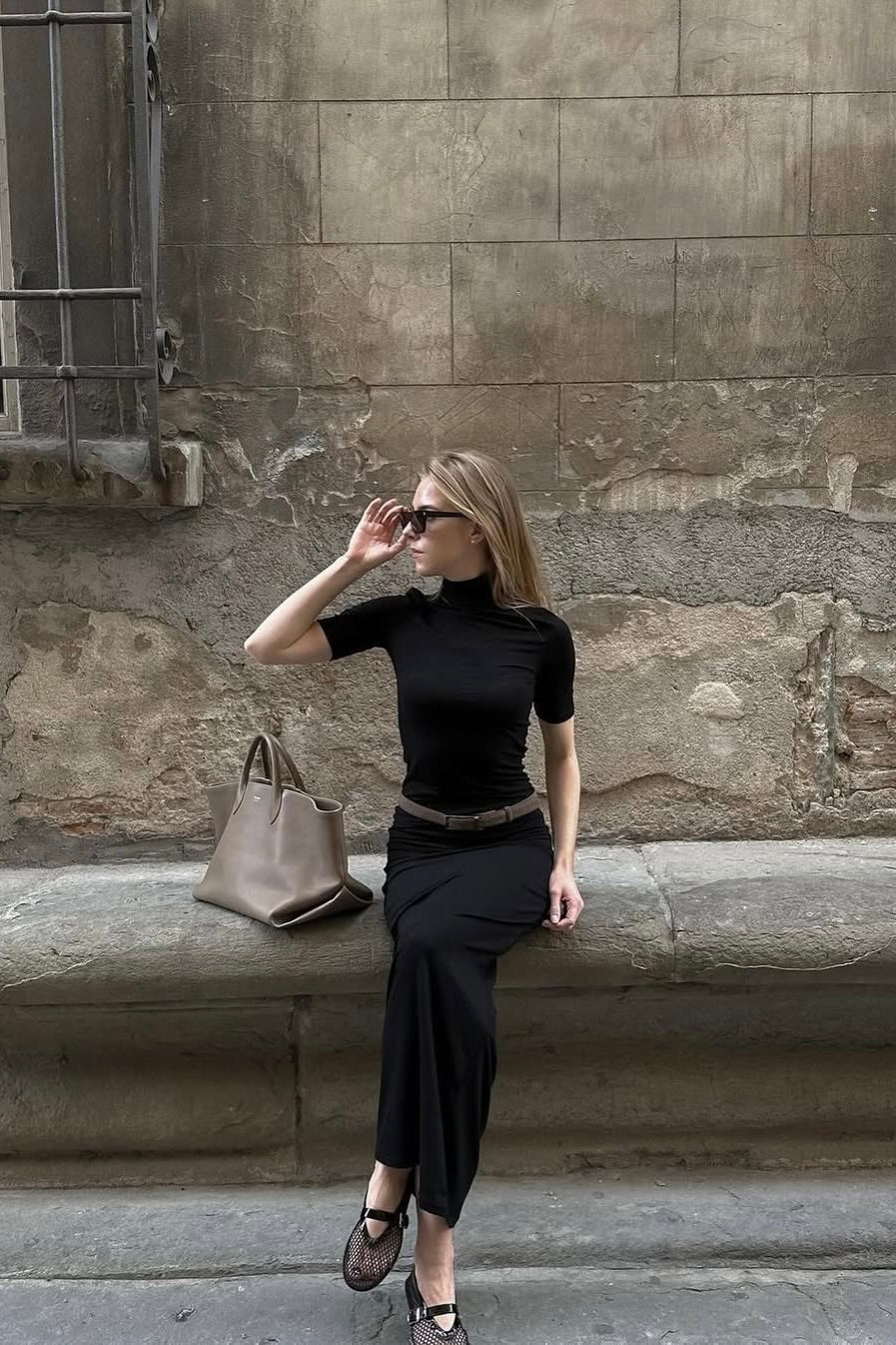 Fashionable woman in a black dress and sunglasses seated on a stone bench, styled for a daytime look.